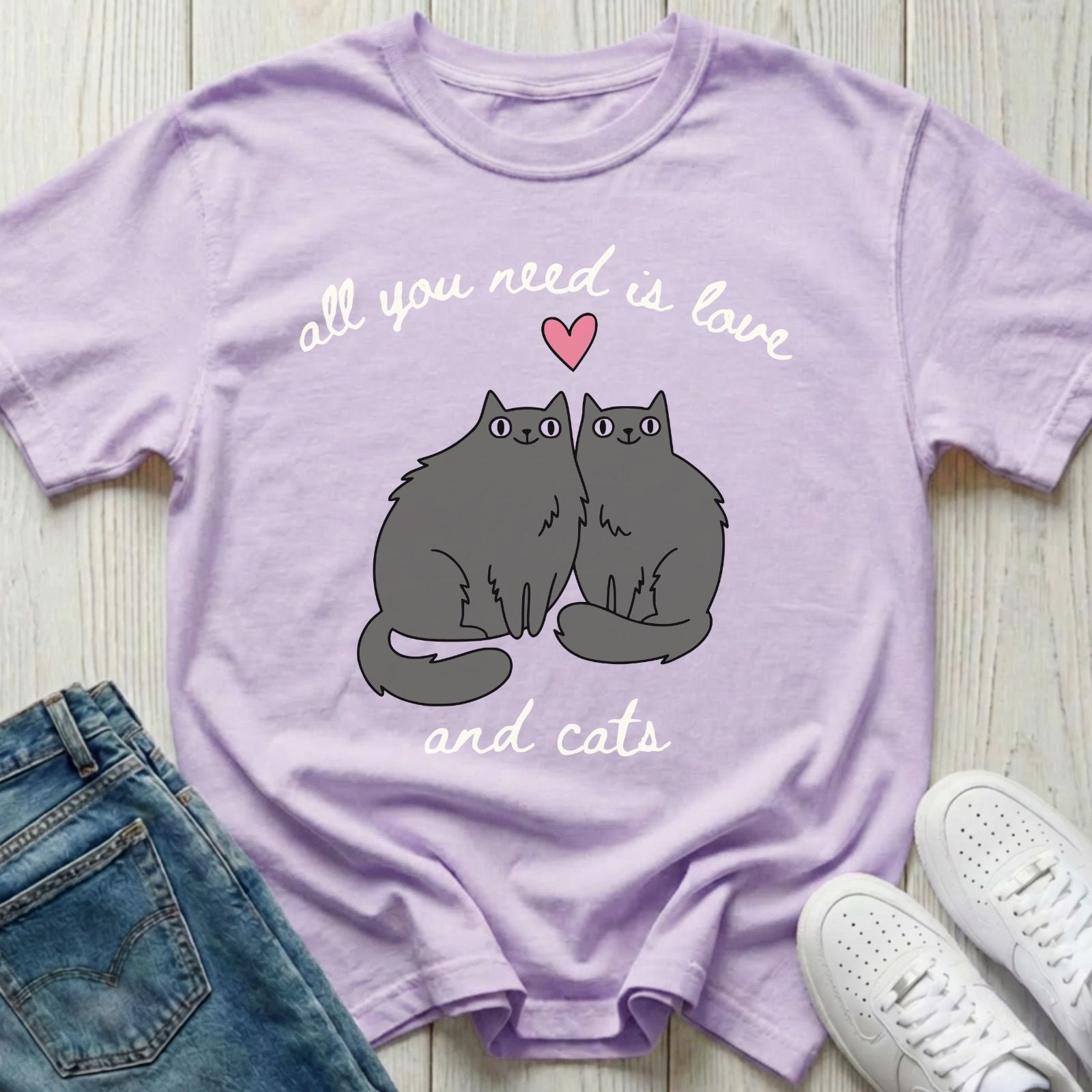 Purple t-shirt with cat design and text 'all you need is love and cats' on a wooden surface with jeans and sneakers.